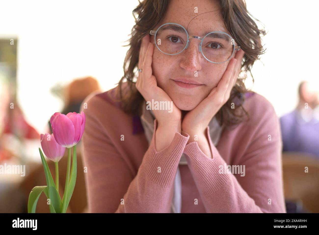 Donna sorridente con gli occhiali che appoggiano il mento sulle mani vicino a un tulipano rosa Foto Stock