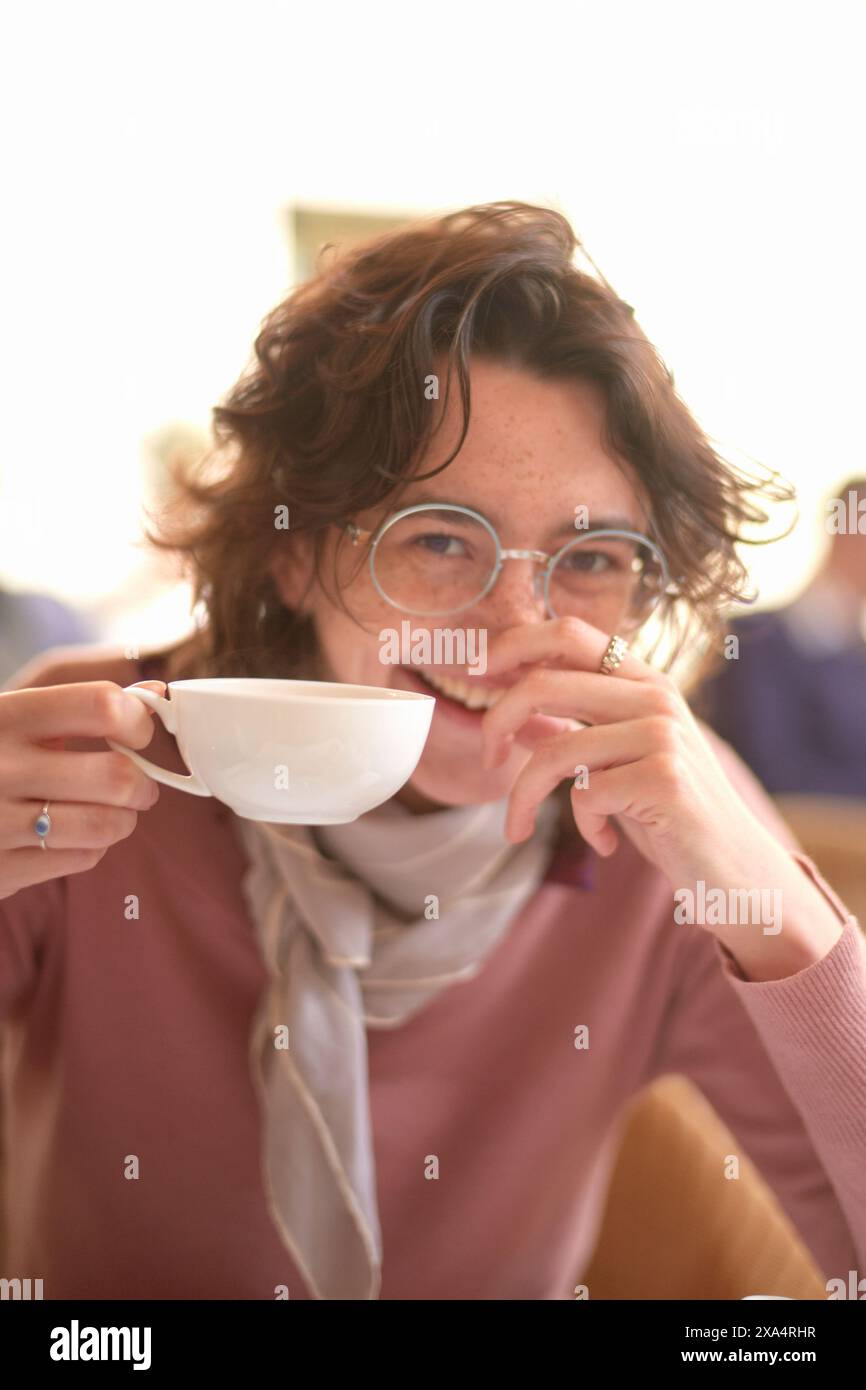 Donna sorridente con bicchieri sorseggiando da una grande tazza bianca in un accogliente ambiente da caffe'. Foto Stock