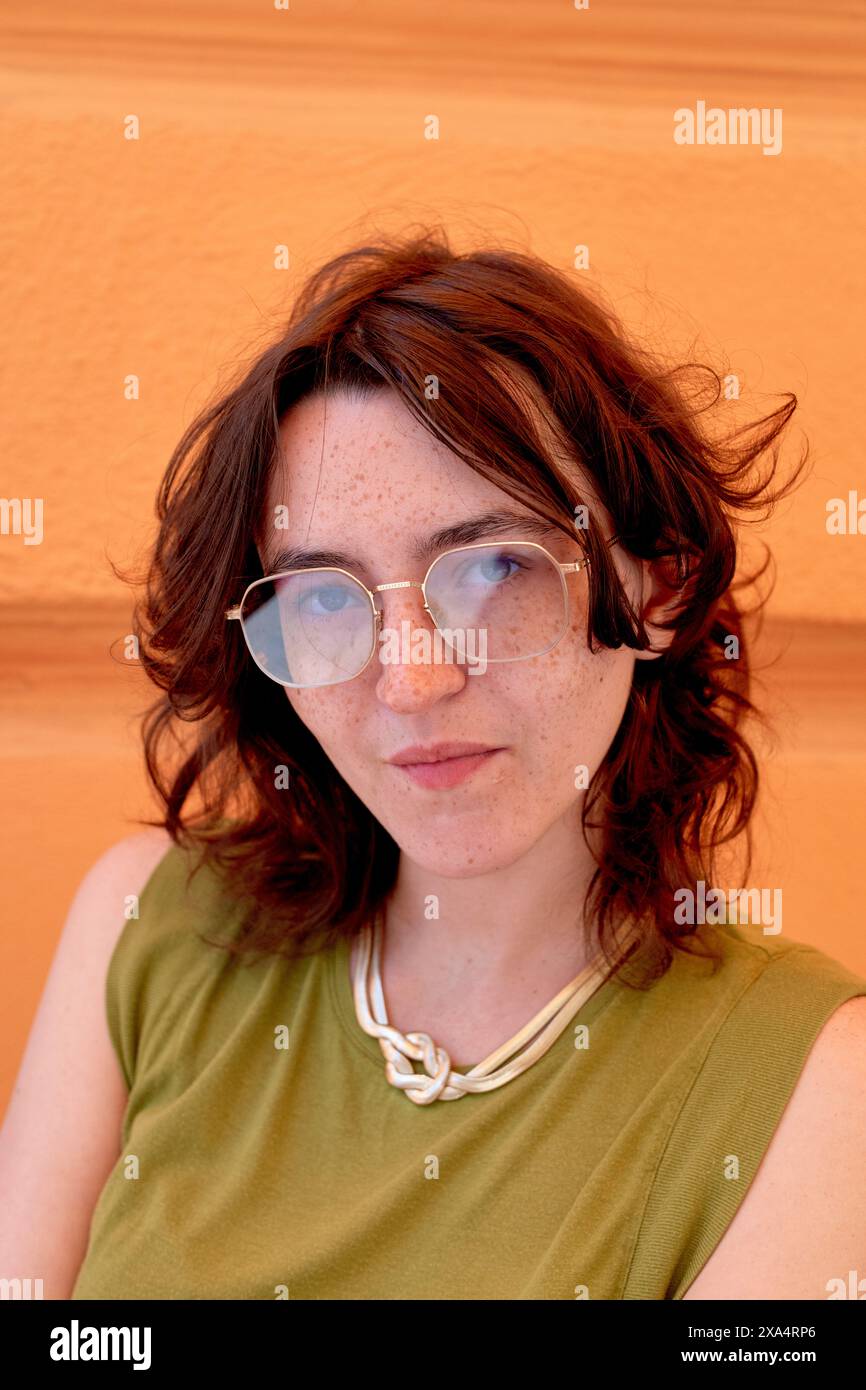 Ritratto informale di una giovane donna lentigginata con i capelli marroni contro una parete arancione e occhiali e un top verde. Foto Stock