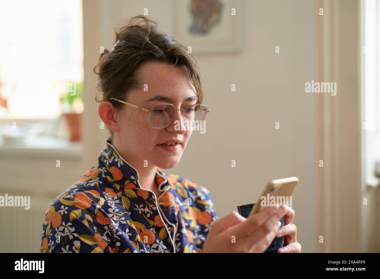 Giovane adulto con occhiali concentrati su uno smartphone in una stanza ben illuminata. Foto Stock