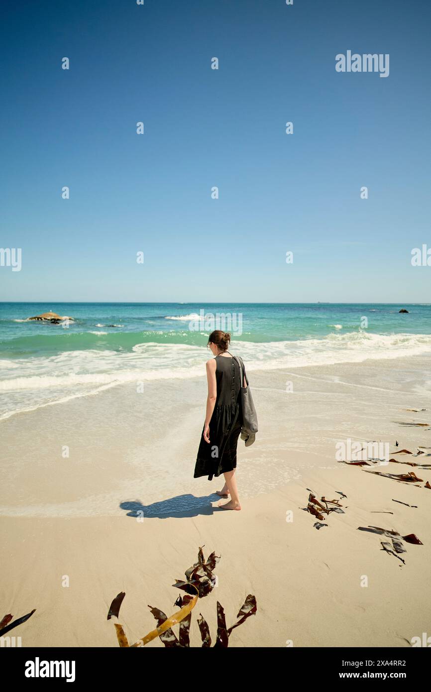 Donna che cammina su una spiaggia sabbiosa vicino all'oceano, porta con sé una borsa, con cieli azzurri. Foto Stock