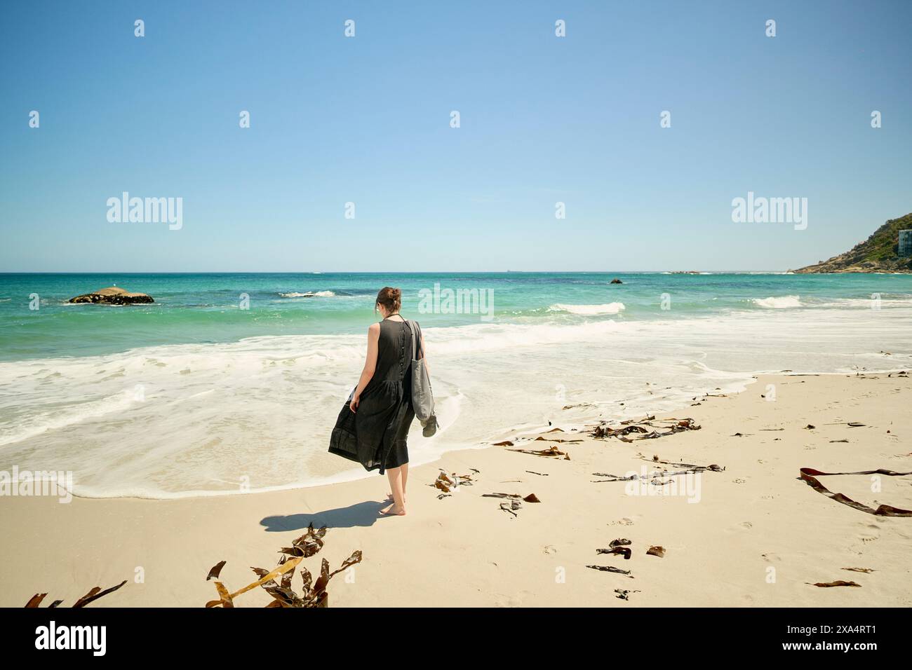 Donna che cammina da sola su una spiaggia soleggiata con onde sullo sfondo e alghe disseminate sulla sabbia. Foto Stock