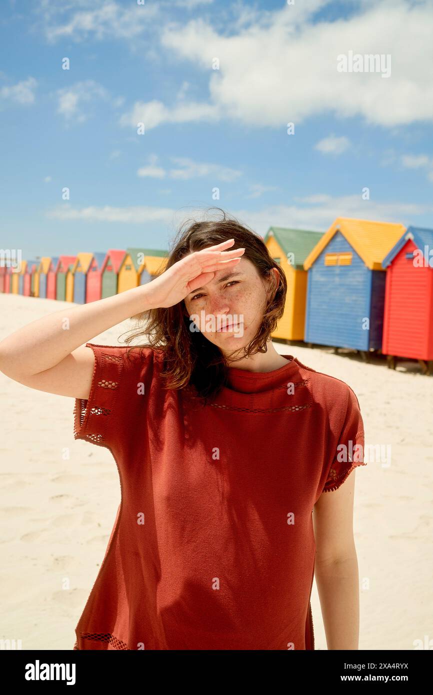 Donna che protegge gli occhi dal sole su una spiaggia sabbiosa con rifugi colorate sullo sfondo. Foto Stock