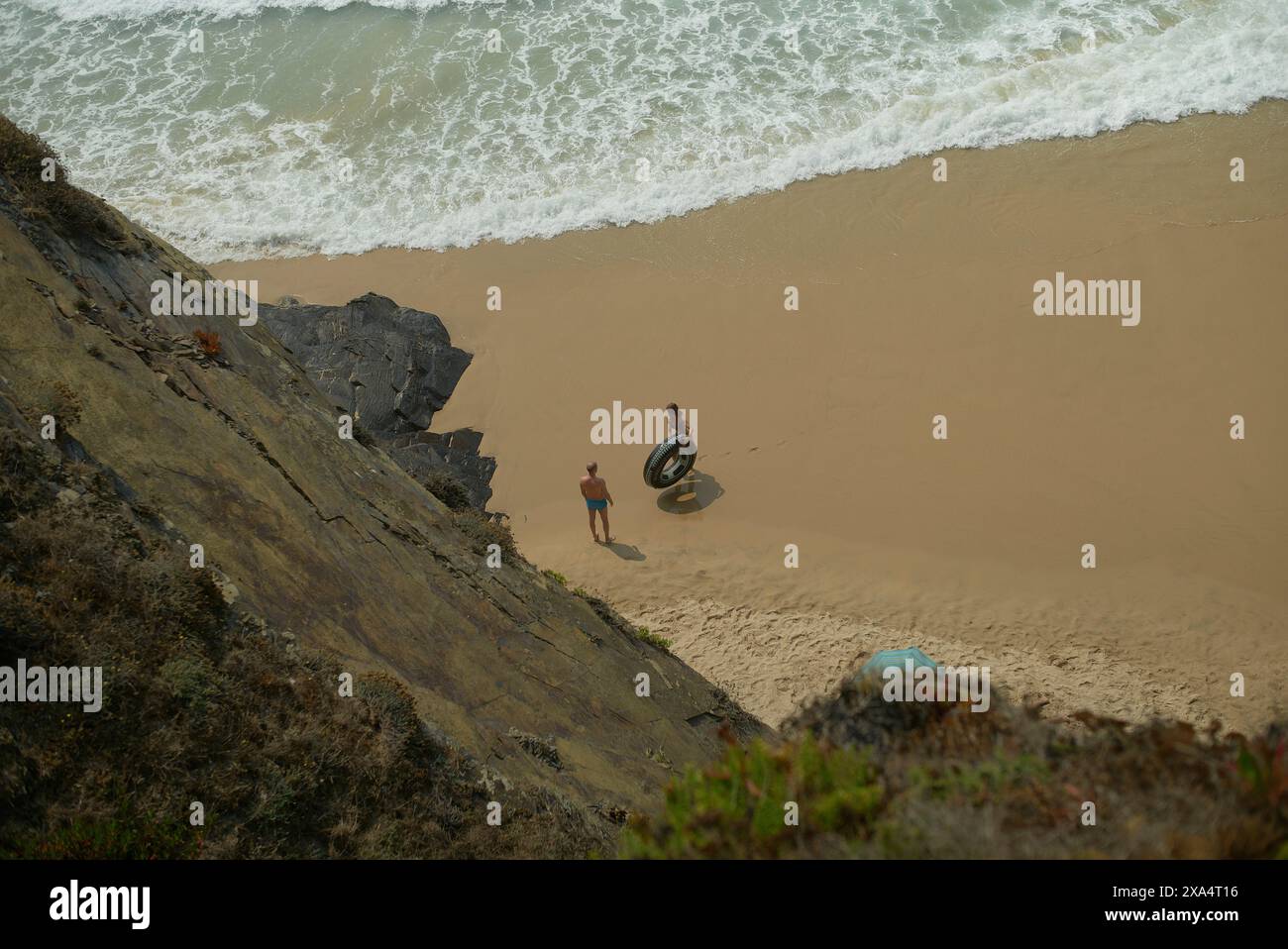 Due persone camminano su una spiaggia sabbiosa accanto a una scogliera con onde che si infrangono sullo sfondo. Foto Stock