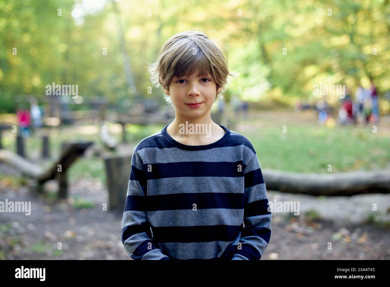 Un ragazzo con un sorriso sottile si erge in un parco soleggiato con una camicia a righe e alberi con foglie autunnali sullo sfondo. Foto Stock