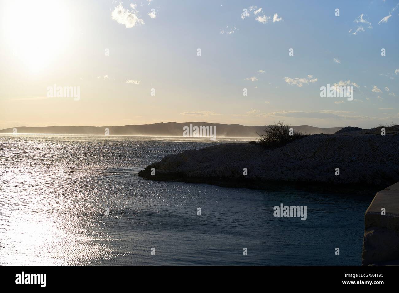 Tramonta un caldo bagliore su un mare tranquillo con montagne lontane e una costa rocciosa. Foto Stock