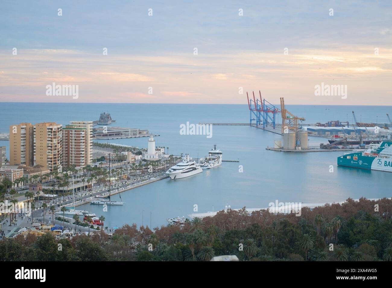 Vista aerea di una città costiera al crepuscolo con un porticciolo con yacht, un porto commerciale con gru ed edifici urbani contro un cielo pastello. Foto Stock