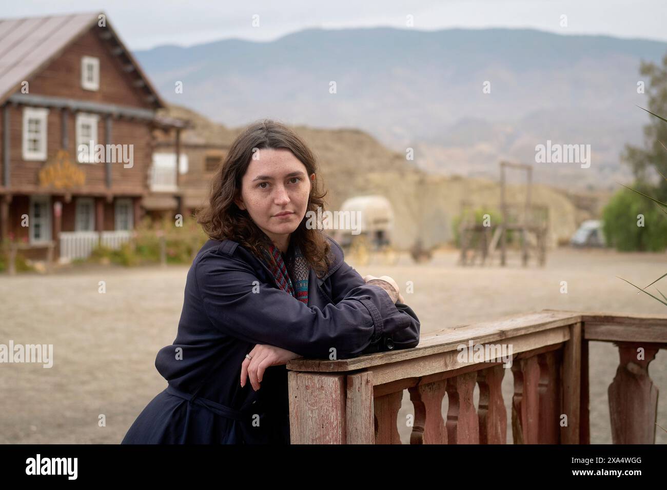 Donna appoggiata su una recinzione di legno con una casa rustica e montagne sullo sfondo. Foto Stock