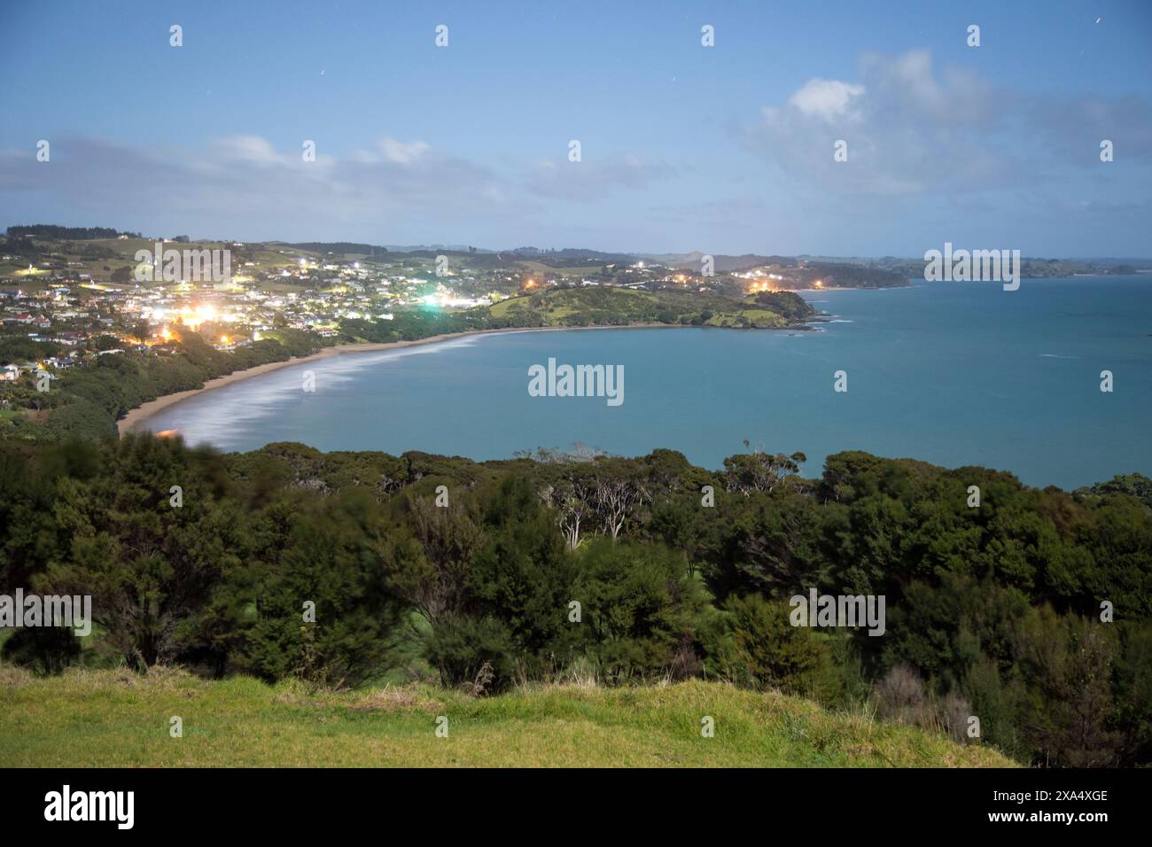 Vista notturna di una città costiera con strade illuminate adiacenti a una baia tranquilla, circondata da colline scure e da un cielo punteggiato di stelle. Foto Stock