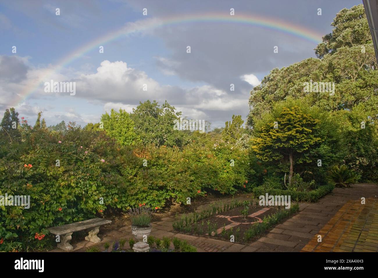 Vibrante arcobaleno che si innalza su un tranquillo giardino con vegetazione lussureggiante e un cielo azzurro limpido dopo le precipitazioni. Foto Stock