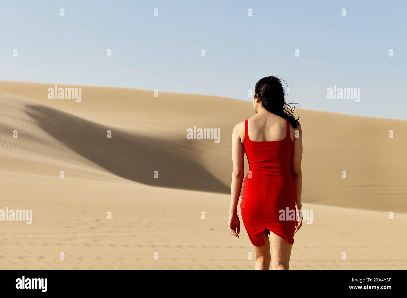 Una donna con un vestito rosso cammina da sola attraverso un paesaggio desertico liscio sotto un cielo limpido. Foto Stock