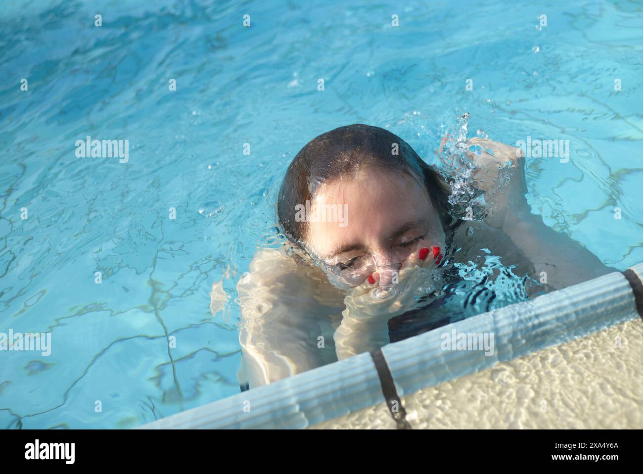 Una persona emerge dall'acqua cristallina della piscina, afferrando il bordo con una mano mostrando lo smalto rosso. Foto Stock