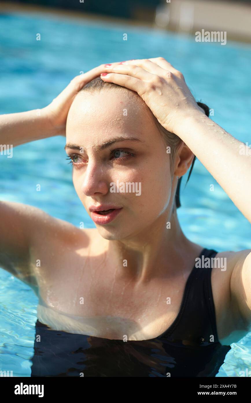 Una donna con un costume da bagno nero emerge da una piscina, brillando sull'acqua sulla pelle, mentre spinge i capelli bagnati indietro con entrambe le mani. Foto Stock