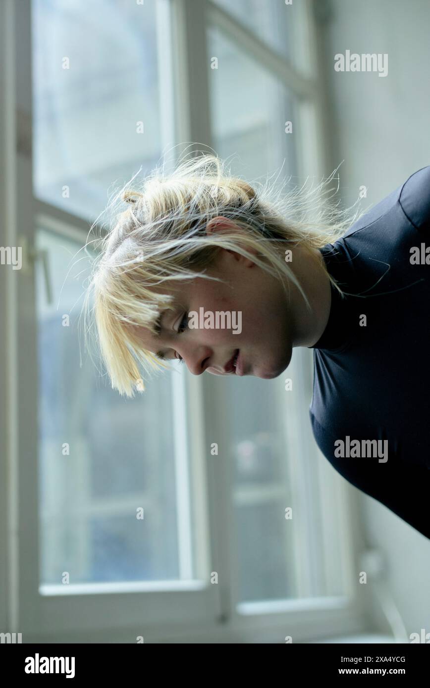 Una persona con capelli biondi in una parte superiore blu che guarda fuori da una finestra con un'espressione premurosa. Foto Stock