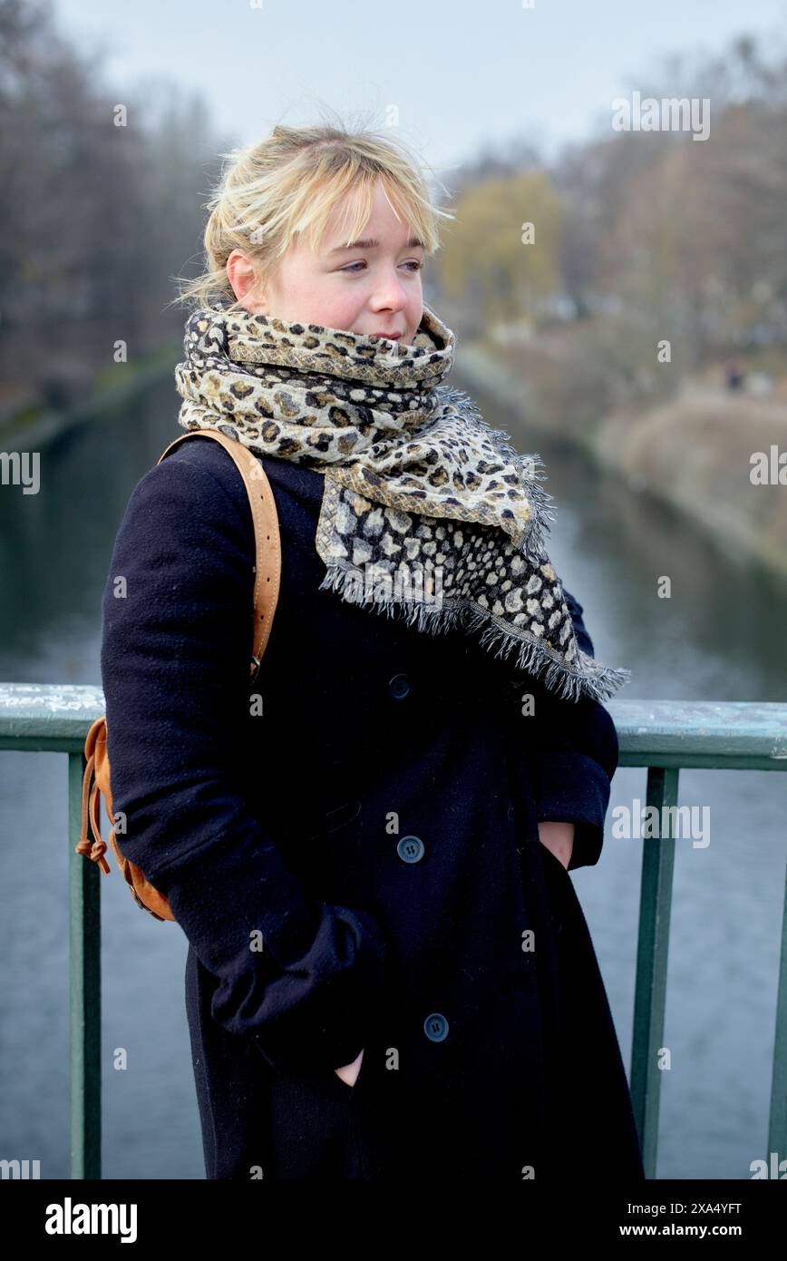 Una donna con un cappotto scuro e una sciarpa a motivi geometrici si erge su un ponte sul fiume, guardando di lato in una giornata nebbiosa. Foto Stock