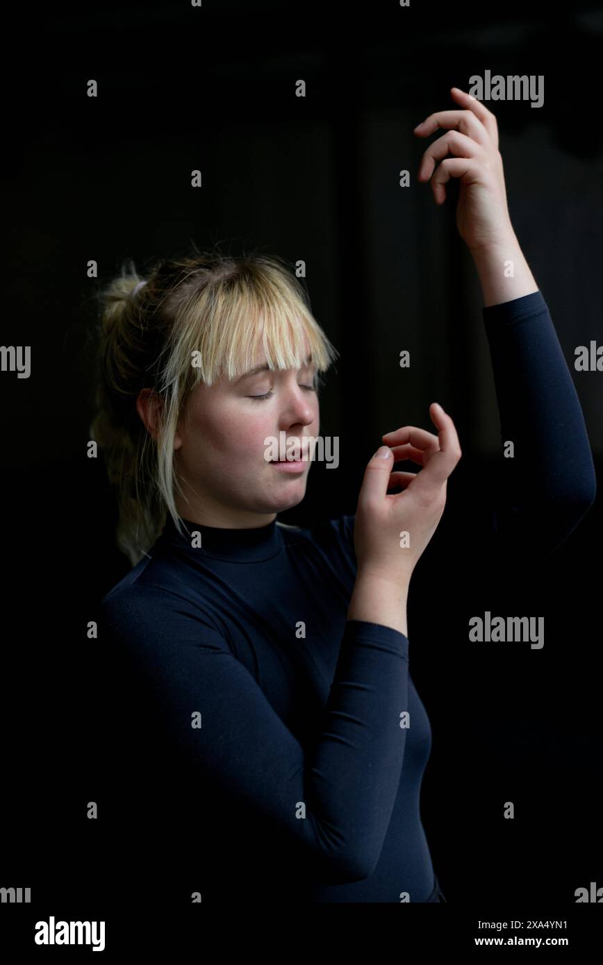 Una giovane donna con gli occhi chiusi, alzando la mano con grazia su uno sfondo scuro in una posa contemplativa. Foto Stock