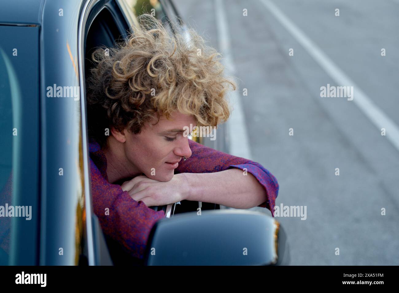 Uomo dai capelli ricci che si sporge dal finestrino dell'auto, godendosi la brezza. Foto Stock