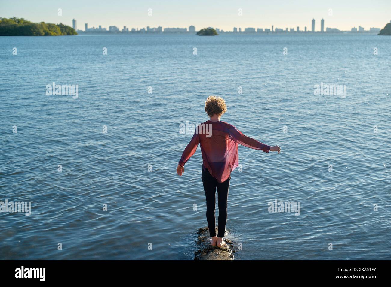 Una persona sta su un molo che si estende le braccia oltre una tranquilla distesa d'acqua con uno skyline della città in lontananza. Foto Stock