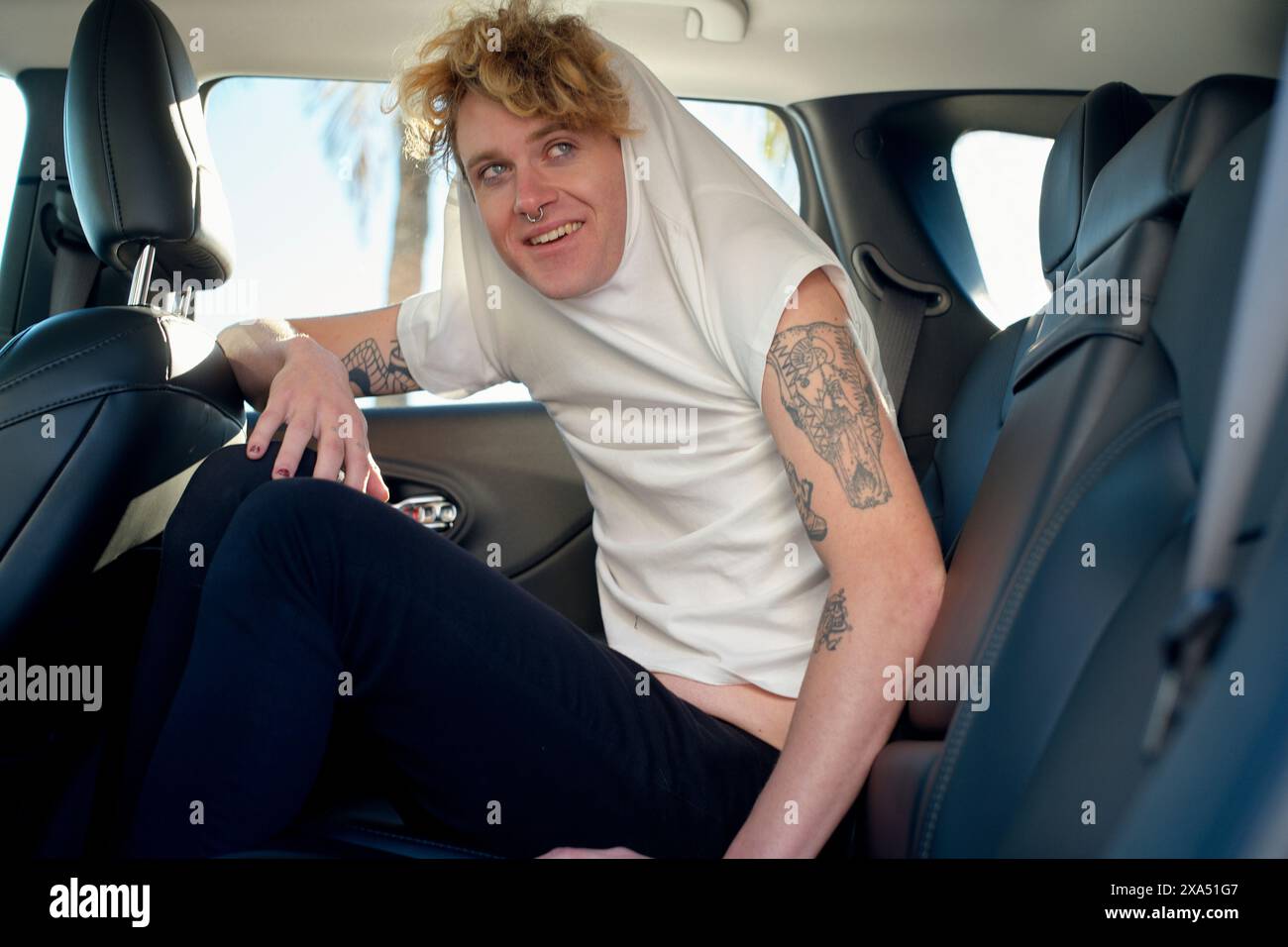 Giovane sorridente con tatuaggi seduto sul sedile posteriore di un'auto, con una felpa con cappuccio bianca e pantaloni neri. Foto Stock
