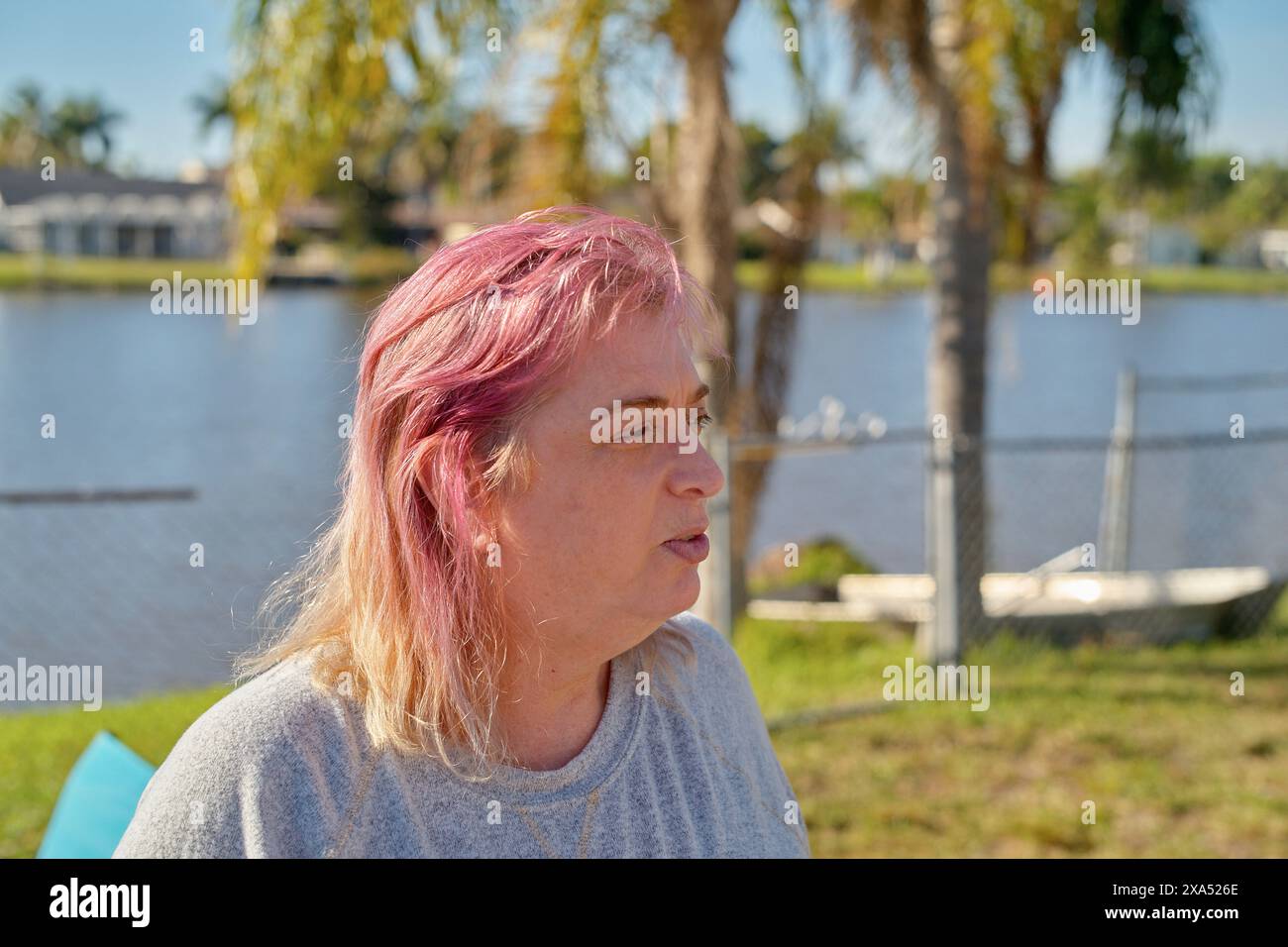 Donna di mezza età con capelli rosa che gode di una giornata di sole in riva al lago con palme sullo sfondo. Foto Stock