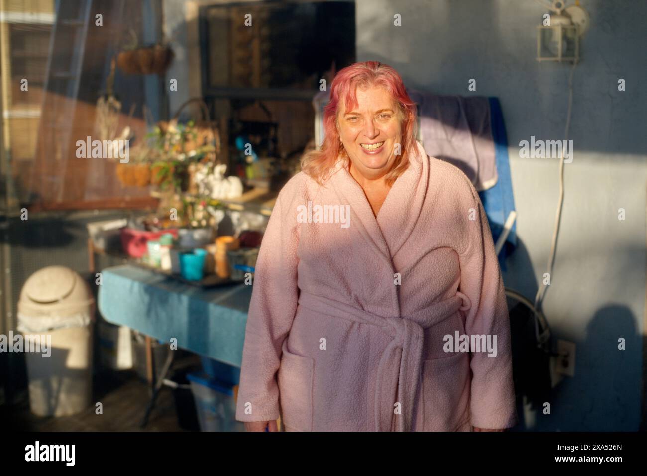 Donna sorridente con i capelli rosa in piedi in una veranda illuminata dal sole e indossa un comodo accappatoio. Foto Stock