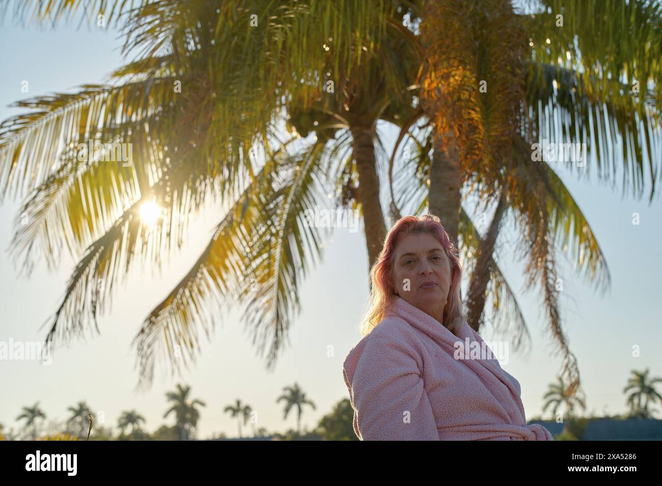 Donna in un accappatoio rosa si trova di fronte alle palme con il sole che tramonta sullo sfondo. Foto Stock