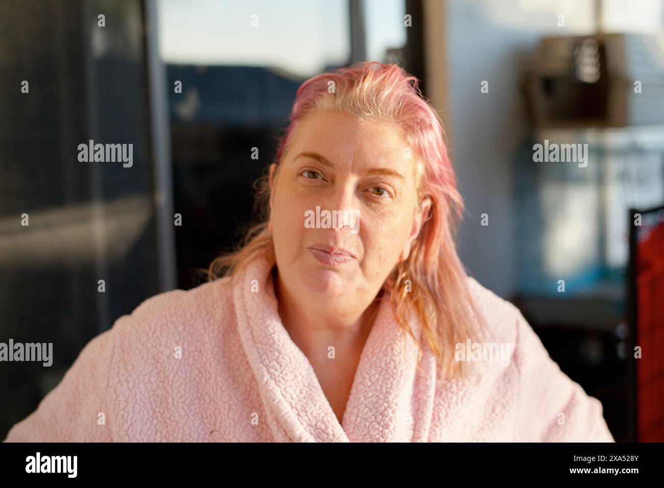 Donna dai capelli rosa avvolto in un comodo accappatoio rosa seduto al chiuso con la luce del sole che illumina il viso Foto Stock