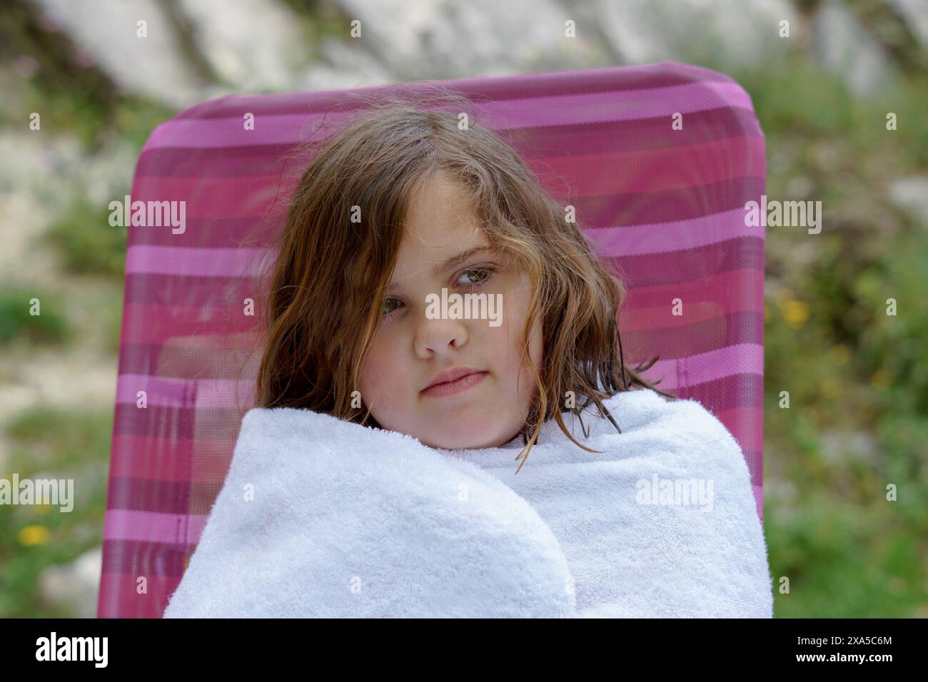 Una bella bambina di otto anni seduta su una sedia avvolta da un asciugamano bianco dopo essere uscita dall'acqua, sembra fredda. Foto Stock