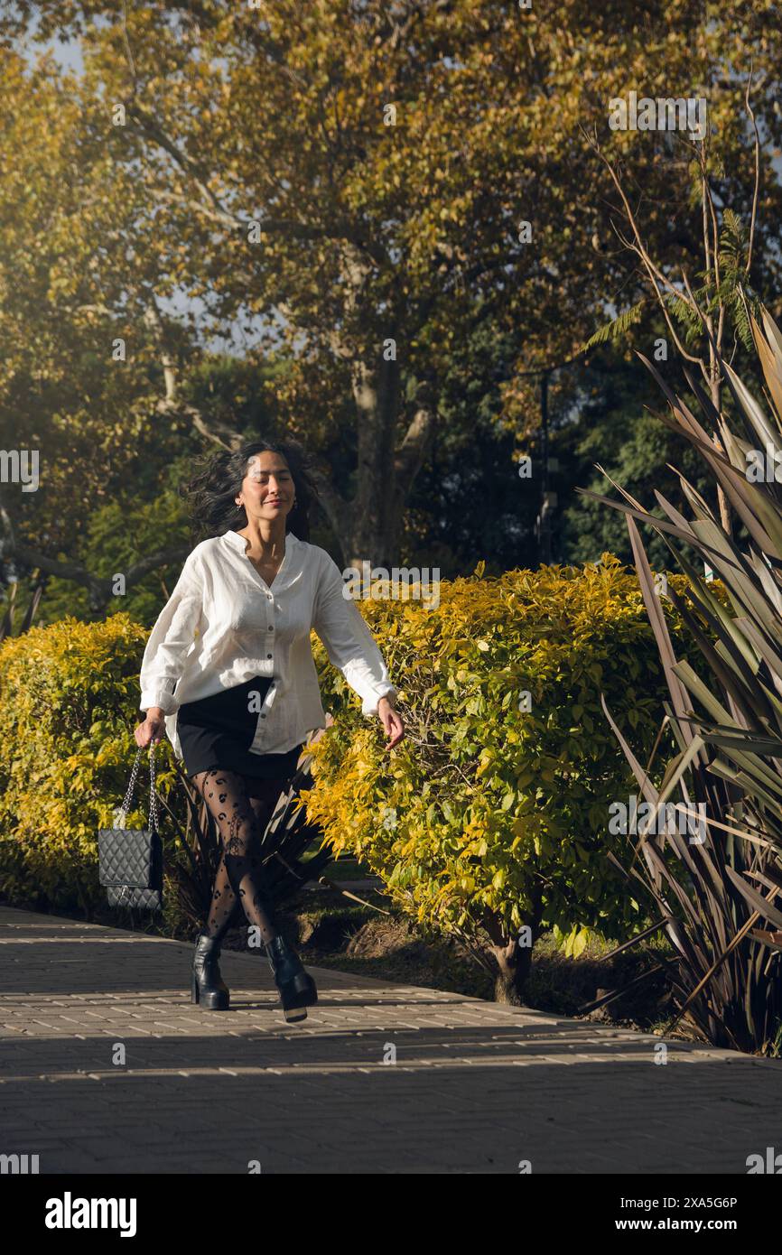 Una donna d'affari sta camminando lungo un sentiero in un parco. Indossa una camicia bianca e pantaloni corti neri con calze collant, cammina in una hu Foto Stock
