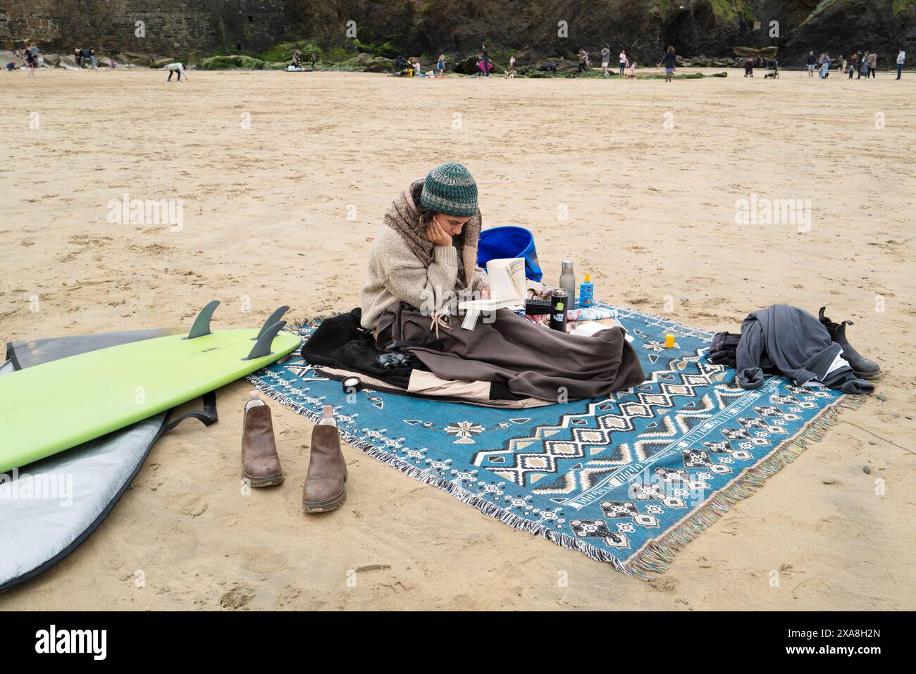Una persona che indossa abiti caldi seduta su un tappeto che legge un libro sulla spiaggia di Towan a Newquay, in Cornovaglia, nel Regno Unito. Foto Stock