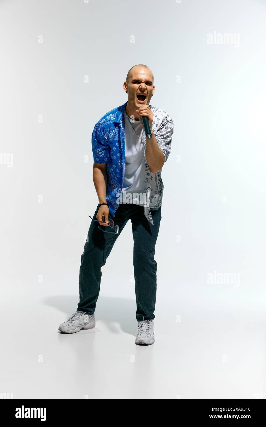 Abbigliamento casual da uomo completo di occhiali, si esibisce energicamente mentre canta in un microfono sullo sfondo bianco dello studio. Foto Stock