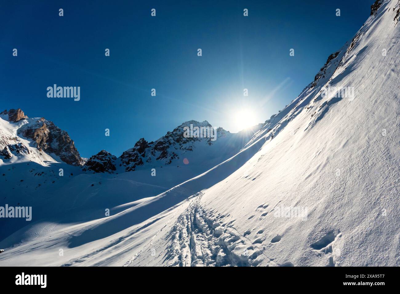 Paesaggio di una montagna ghiacciata con il sole che tramonta dietro le cime rocciose - un sentiero di alpinismo che si snoda su terreni innevati Foto Stock