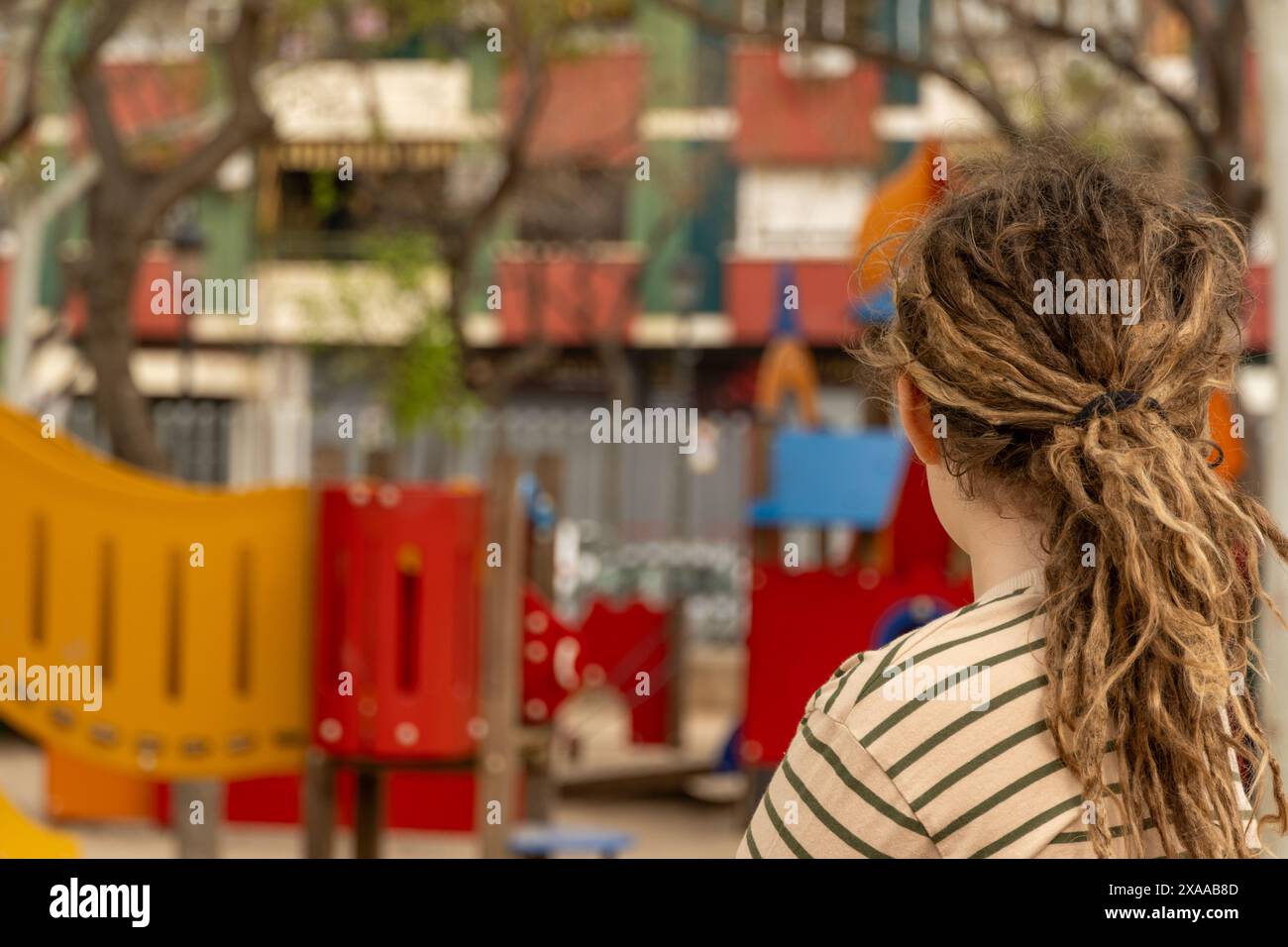 un ragazzo dai capelli biondi con dreadlocks, che osserva il vivace parco giochi da una prospettiva unica. Innocenza e avventura si intrecciano mentre il bambino si diverte Foto Stock