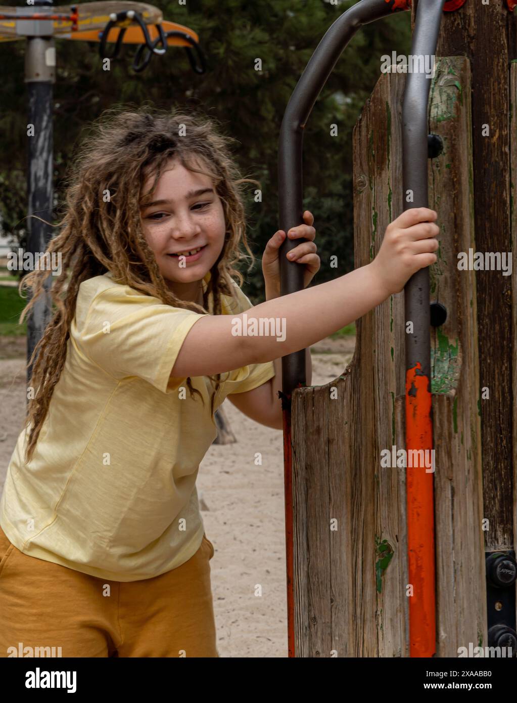 Esplora l'infanzia radiosa in questa immagine verticale: Un ragazzo con lunghi capelli biondi, con dreadlocks, gode di una giornata calda al parco giochi. Il suo sma infettivo Foto Stock