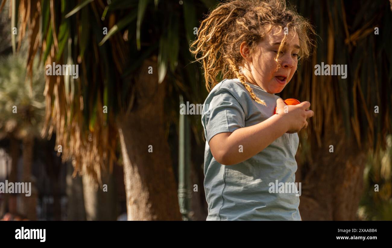 un ragazzo biondo con i dreadlock sorride al sole in un parco alberato. La natura brilla mentre la felicità infantile illumina la scena, creando un momento pieno Foto Stock