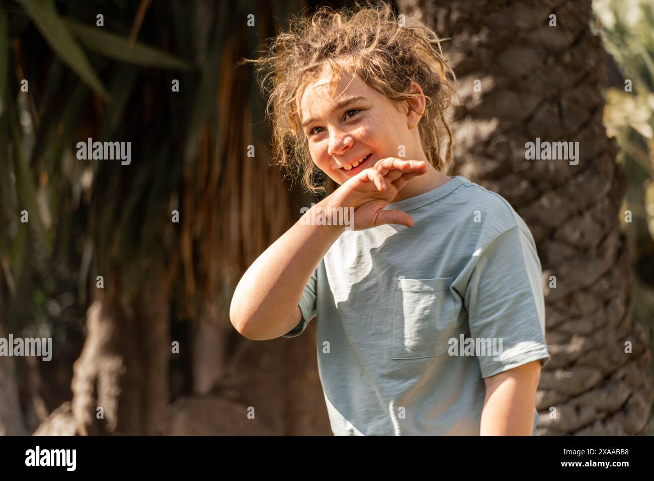 il ragazzo biondo con i dreadlocks sorride in un parco alberato sotto il sole. Una scena affascinante che evoca l'infanzia, la gioia e il legame con la natura Foto Stock