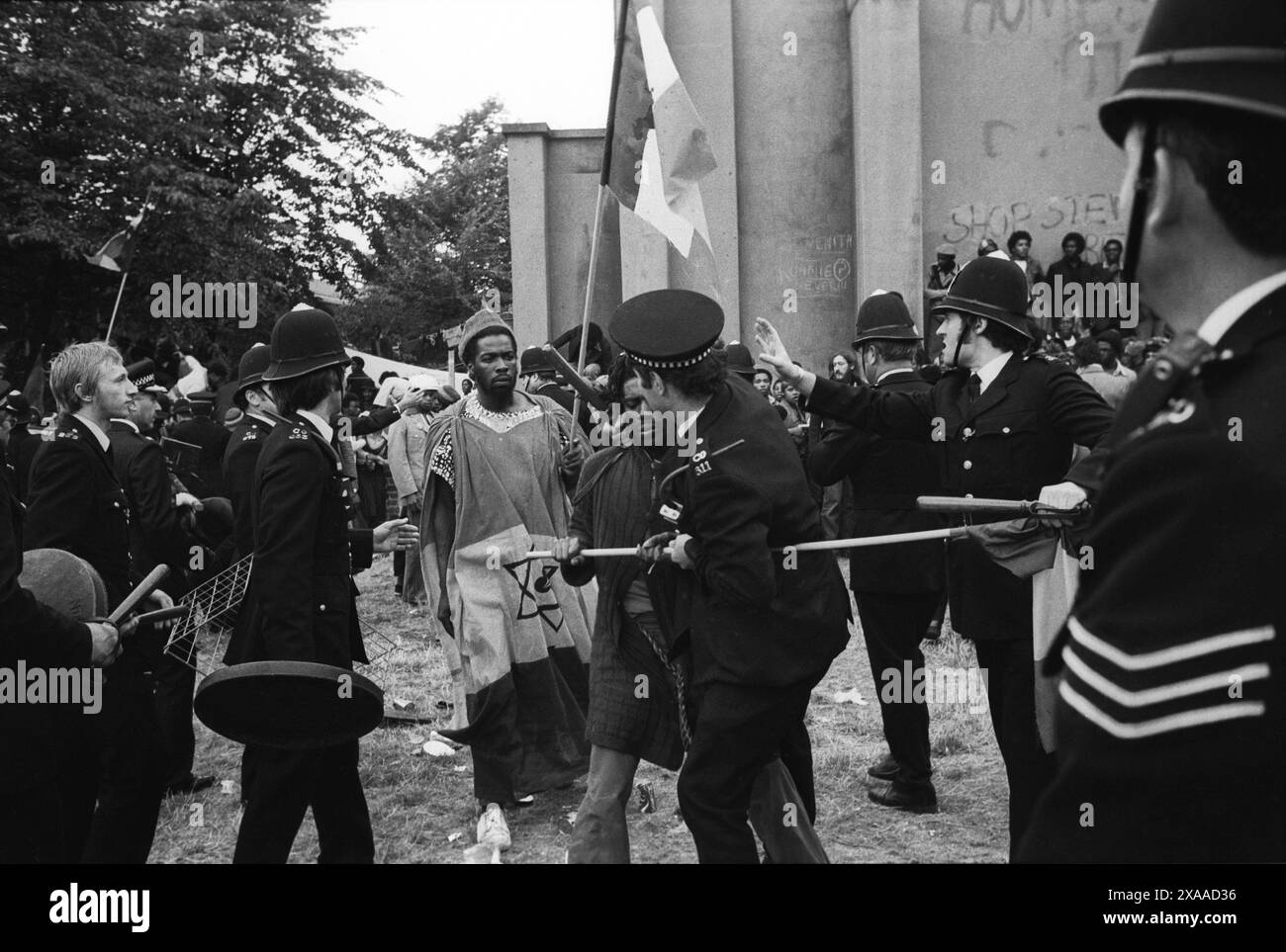 La rivolta del carnevale di Notting Hill agosto festivi lunedì 1976. Un gruppo di Rastafariani britannici neri che cantano e suonano tranquillamente. La polizia ha rotto il gruppo pensando che fossero dei rivoltosi. Alcuni poliziotti trasportano coperchi per la pattumiera da usare come scudo e hanno disegnato i loro manichini. Notting Hill, Londra, Inghilterra 30 agosto. 1970 UK HOMER SYKES Foto Stock