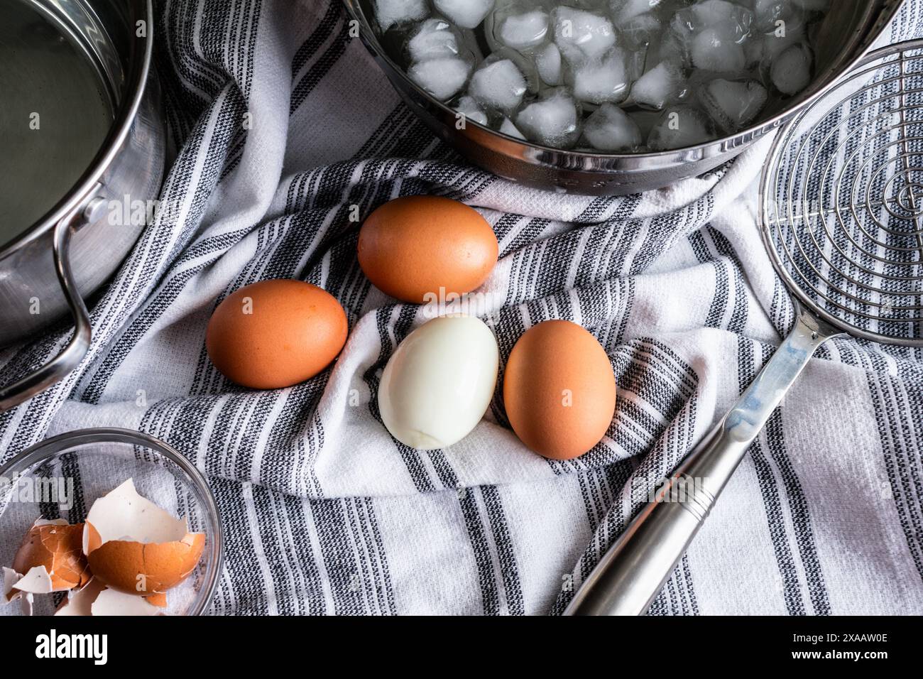 Uova sode intere pelate e non sbucciate: Quattro uova sode su un asciugamano da cucina vicino a una ciotola di acqua ghiacciata e uno skimmer di metallo Foto Stock