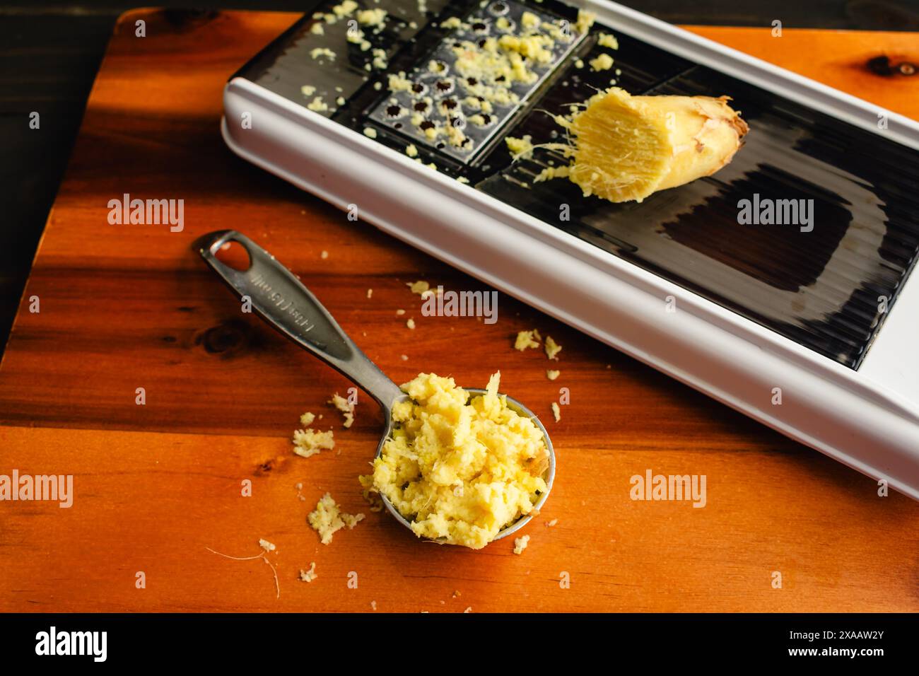 Cucchiaio di zenzero grattugiato con una grattugia: Un misurino di zenzero grattugiato su un tagliere di legno con un mandolino Foto Stock
