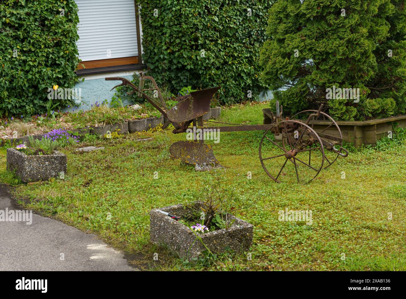 Un vecchio attrezzo agricolo arrugginito è seduto sull'erba di fronte a una casa con arbusti verdi. Foto Stock