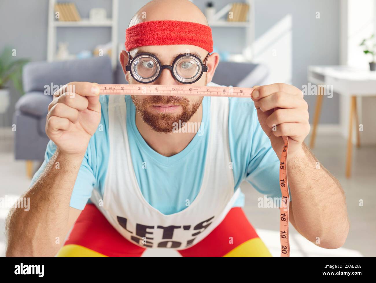 Divertente sportivo con espressione comica effettua misurazioni con il metro a nastro prima di allenarsi a casa Foto Stock