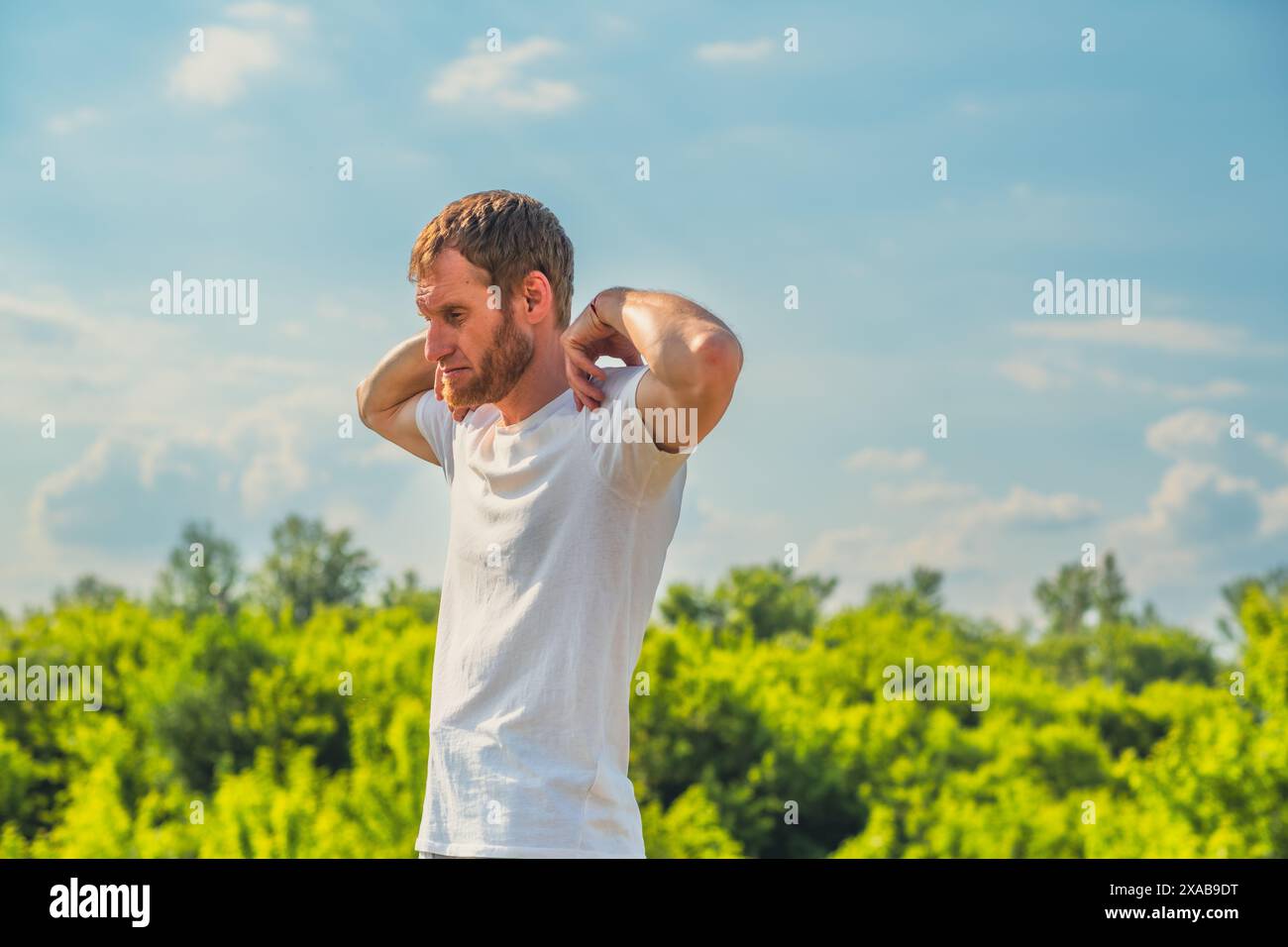 Un uomo con una t-shirt bianca si allunga il braccio mentre è in piedi fuori in una giornata di sole. Sta allungando i muscoli del braccio e della spalla. Foto Stock Un uomo con una t-shirt bianca si allunga il braccio mentre è in piedi fuori in una giornata di sole. Sta allungando i muscoli del braccio e della spalla. Foto Stock