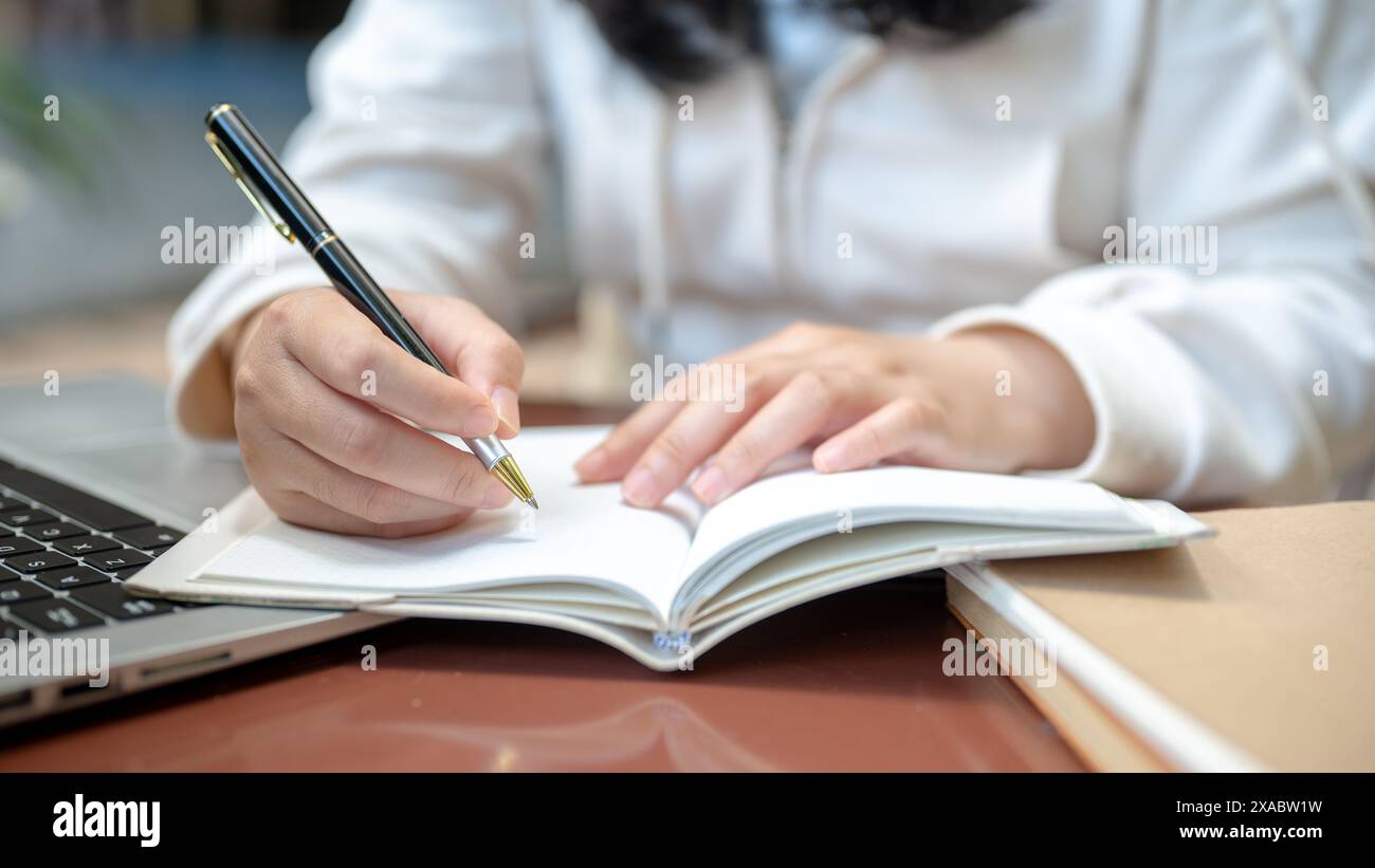 Un'immagine ravvicinata ritagliata di una donna asiatica in abiti casual sta scrivendo qualcosa nel suo taccuino mentre guarda un video sul suo laptop e sul suo ufficio Foto Stock