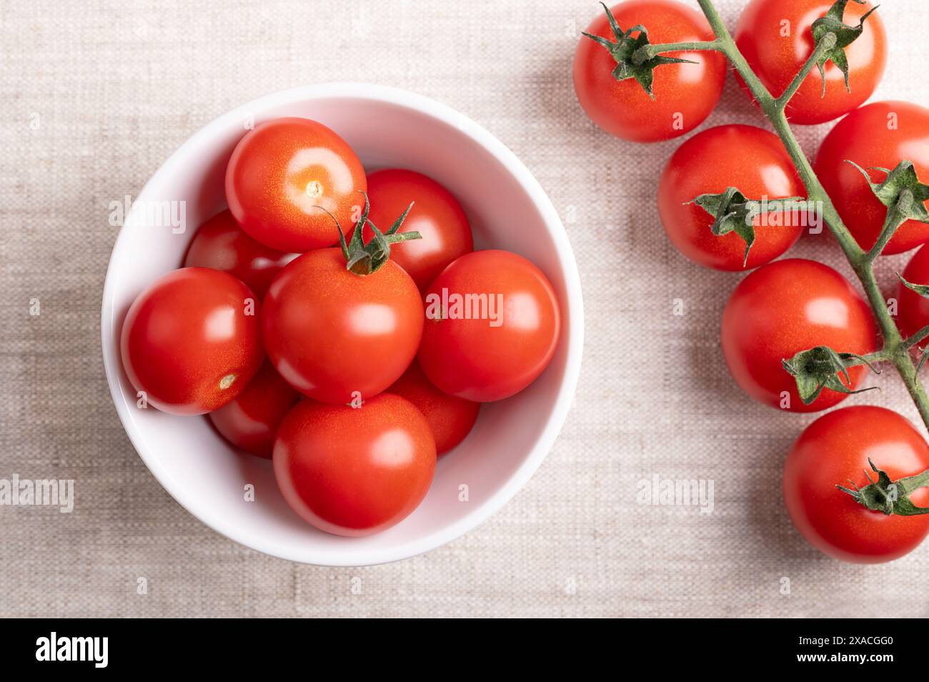 Pomodori ciliegini rossi in una ciotola bianca su tessuto di lino. Pomodori da cocktail piccoli e rotondi freschi e maturi, sul lato sinistro ancora sulla vite. Foto Stock