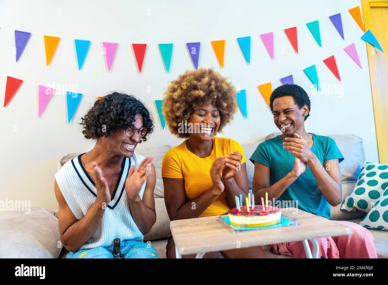 Amici maschi multietnici che si congratulano con una donna africana con una torta di compleanno seduta sul divano sorridendo e applaudendo a casa Foto Stock