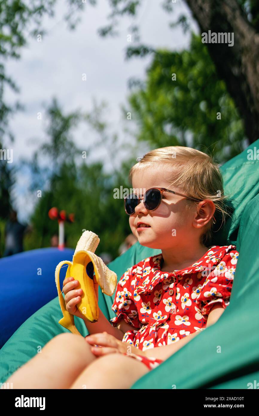 Bambina che mangia una banana. Ritratto di una bella bambina elegante e alla moda con occhiali da sole e un abito rosso mentre si siede su una sedia a sacco Foto Stock