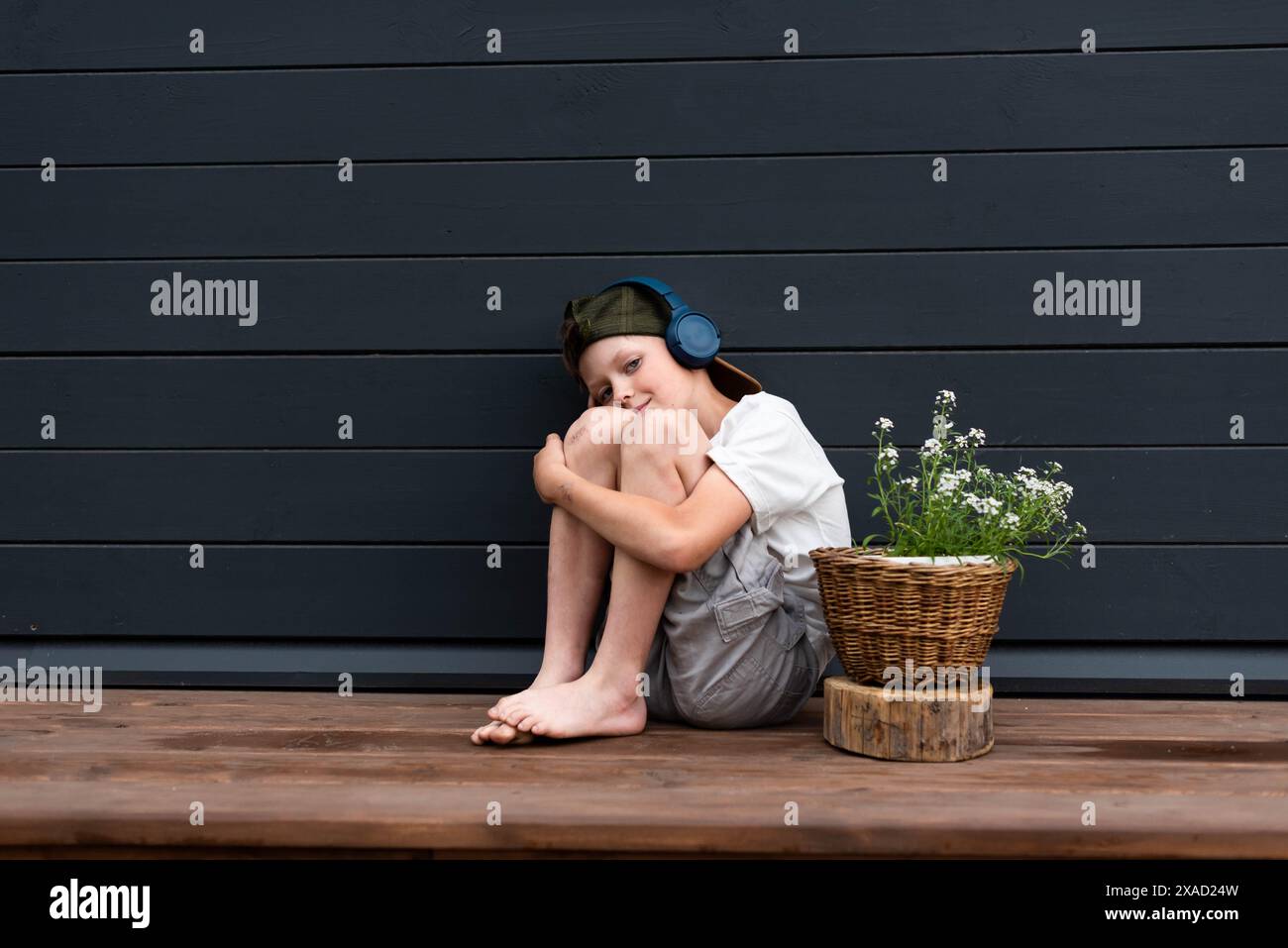 Simpatico ragazzo preadolescente in cuffia che ascolta la musica seduto su una terrazza di legno abbracciando le ginocchia Foto Stock
