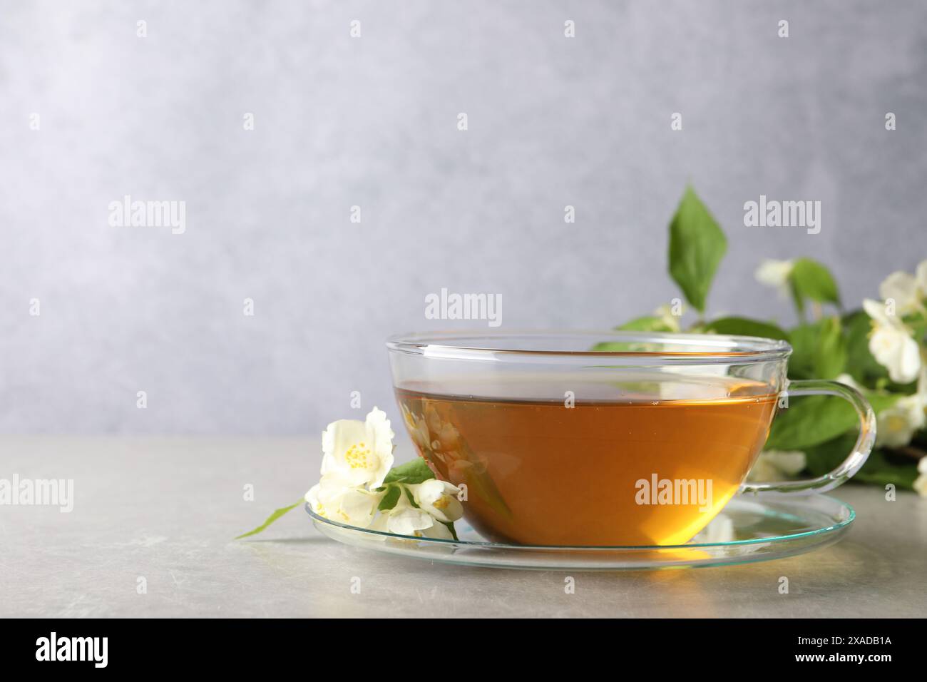 Tè aromatico al gelsomino in tazza, fiori e foglie verdi su un tavolo grigio chiaro, spazio per il testo Foto Stock