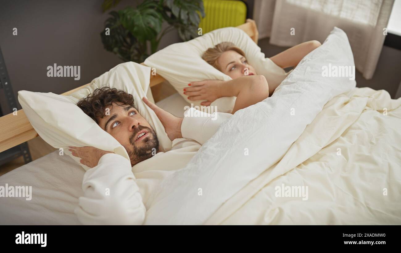 Un uomo e una donna coprono le orecchie con cuscini in una camera da letto interna, raffigurando il rumore di una coppia a casa. Foto Stock
