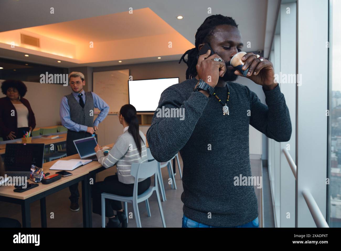 Un uomo nero imprenditore con dreadlocks sta bevendo caffè e ascoltando la telefonata in piedi davanti alla finestra dell'ufficio. Indossa un maglione Foto Stock